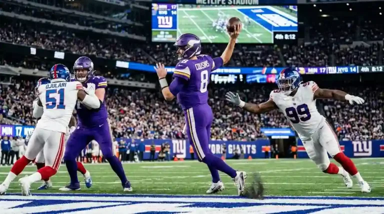 A Minnesota Vikings quarterback in a purple uniform winds up to throw a deep pass while New York Giants defenders close in under bright MetLife Stadium lights.