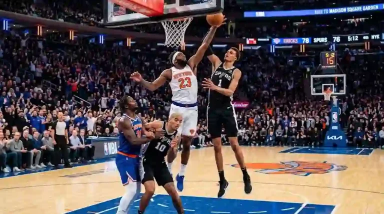 Action-packed NBA game between the New York Knicks and San Antonio Spurs, showing players leaping for a rebound near the basket inside a packed arena.