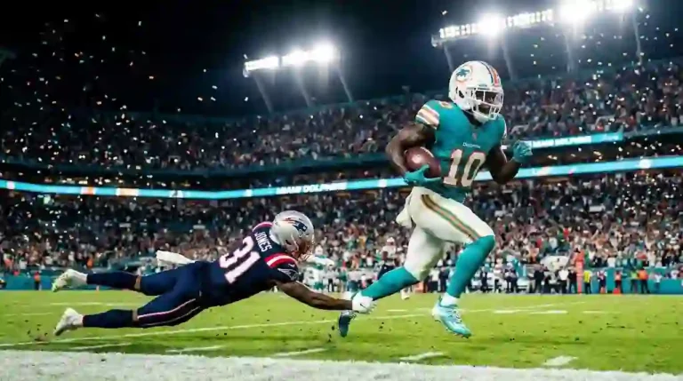 A Miami Dolphins player in aqua and orange uniform sprinting with the football past a diving New England Patriots defender in a packed NFL stadium at night.